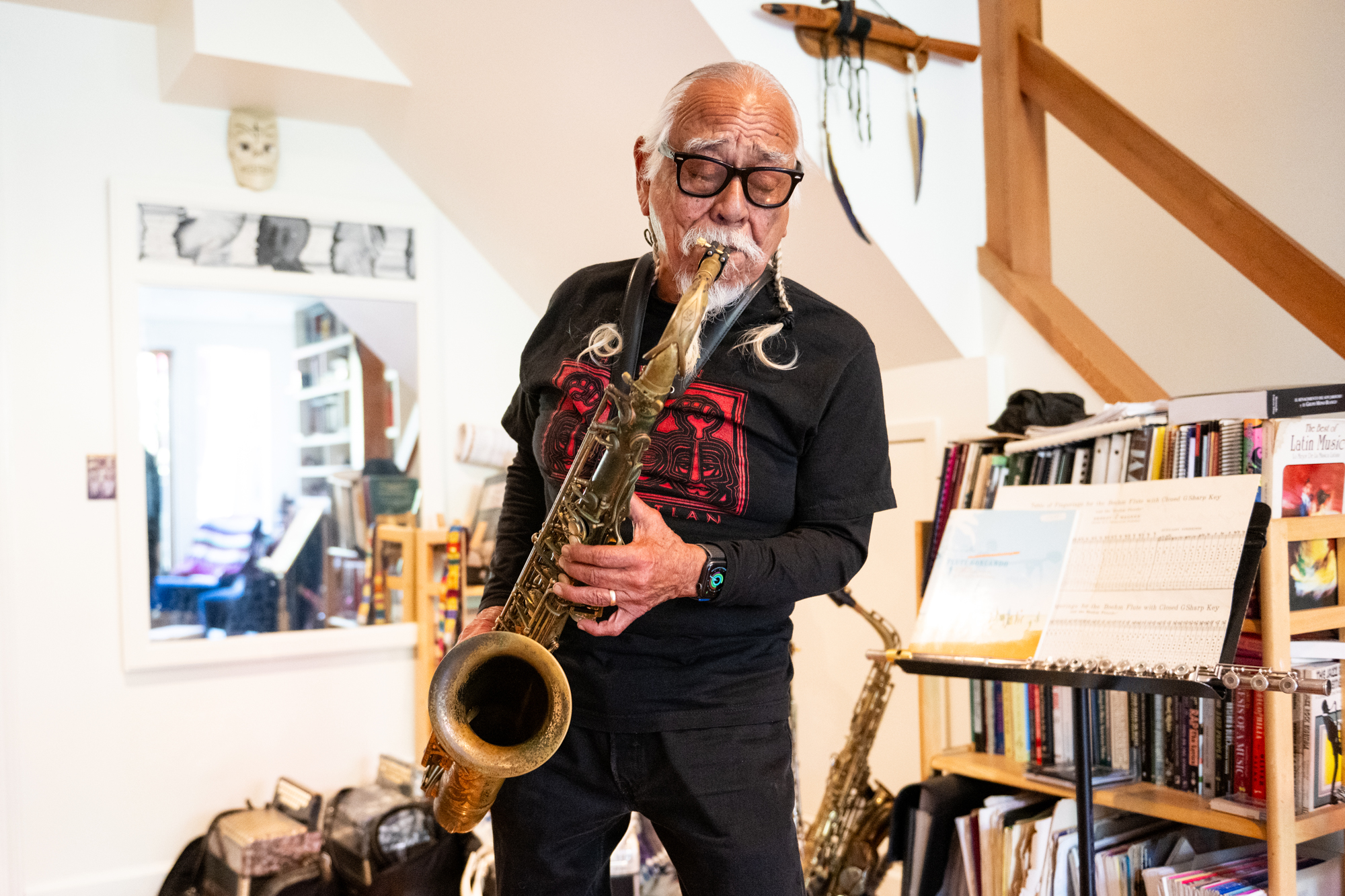 An older man with white braids and glasses plays a saxophone in his home