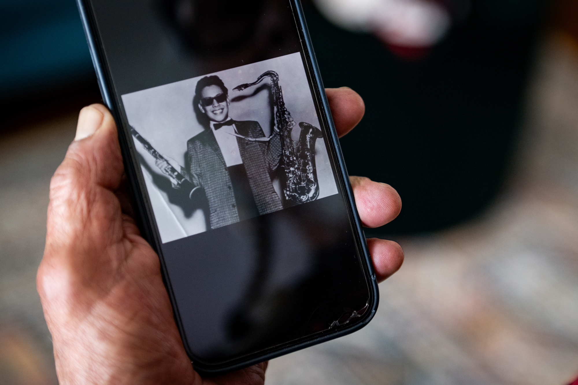 A hand holds a cell phone with a black and white photograph on it of a young man holding a clarinet and a saxophone, wearing a suit and sunglasses