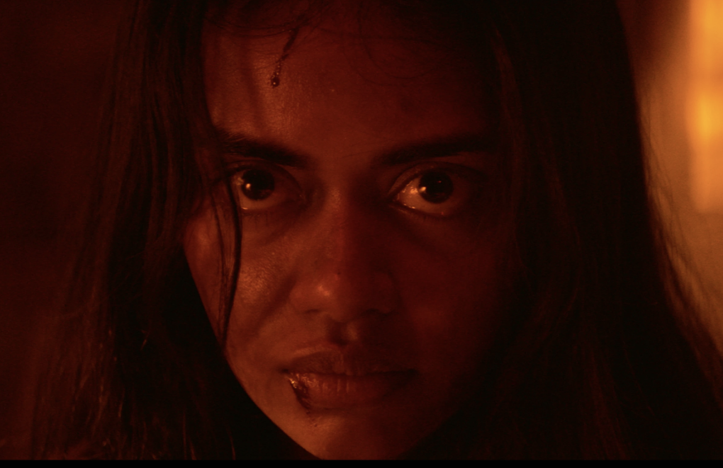 A close up dimly lit shot of a young, menacing looking, wide-eyed South Asian woman.