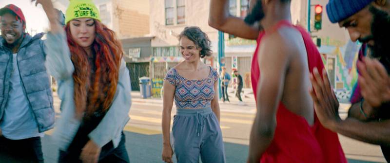 A woman with short brown hair, a leotard and pants smiles at dancers on the street, with storefronts in the background