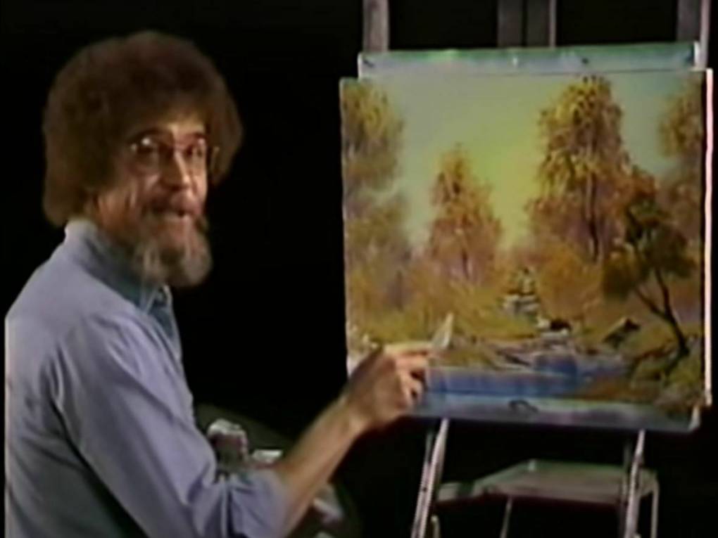 A bearded man with curly hair sits at a canvas, paintbrush in hand. The canvas has a painting of trees and a winding river on it.