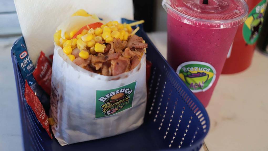 a Brazilian-style burger (which includes corn, bacon, and potato straws) is served in a blue basket with a purple dragonfruit juice besides it