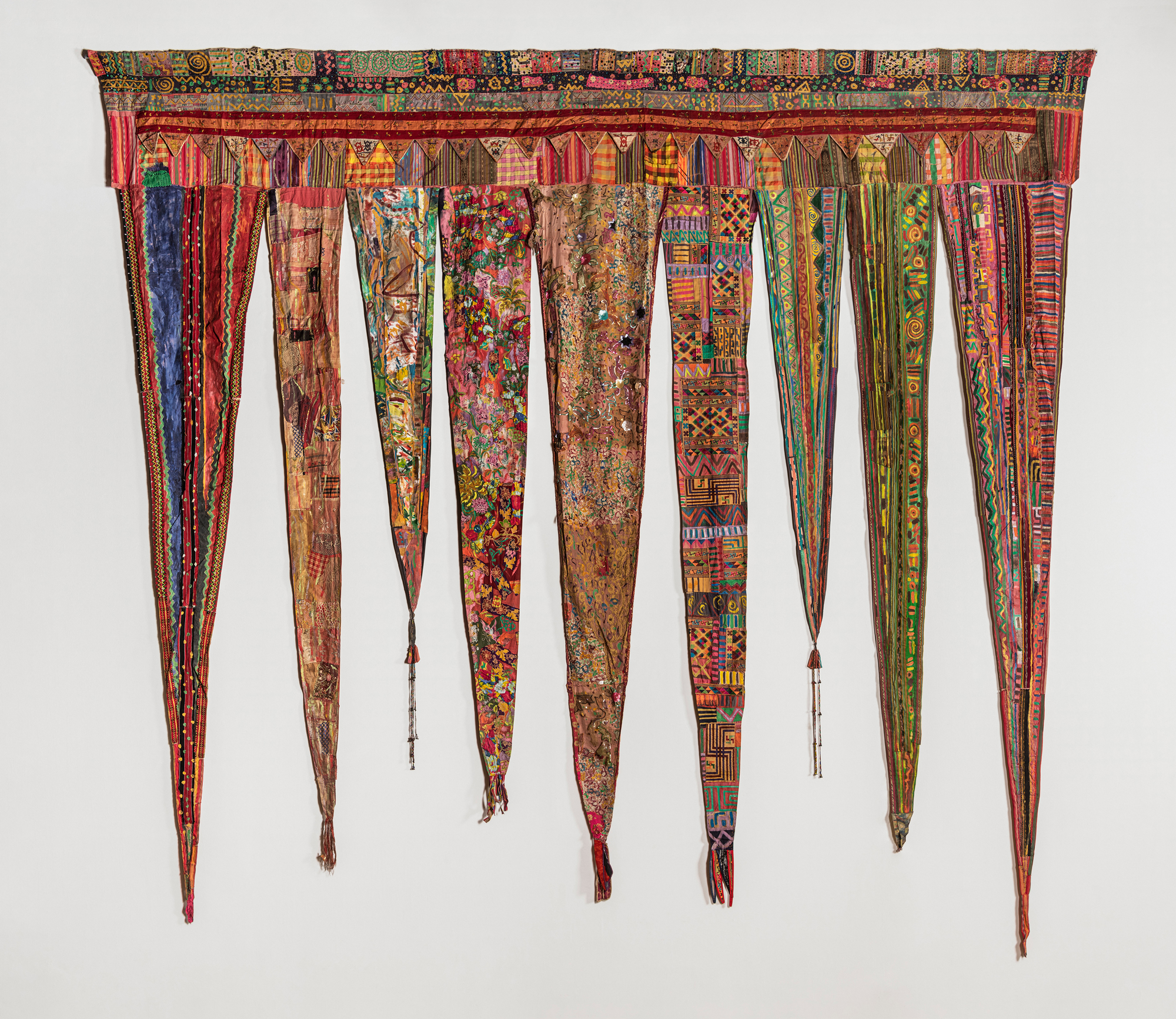 Textile piece made up of many fabrics and patterns, with nine long thin triangles descending from top band