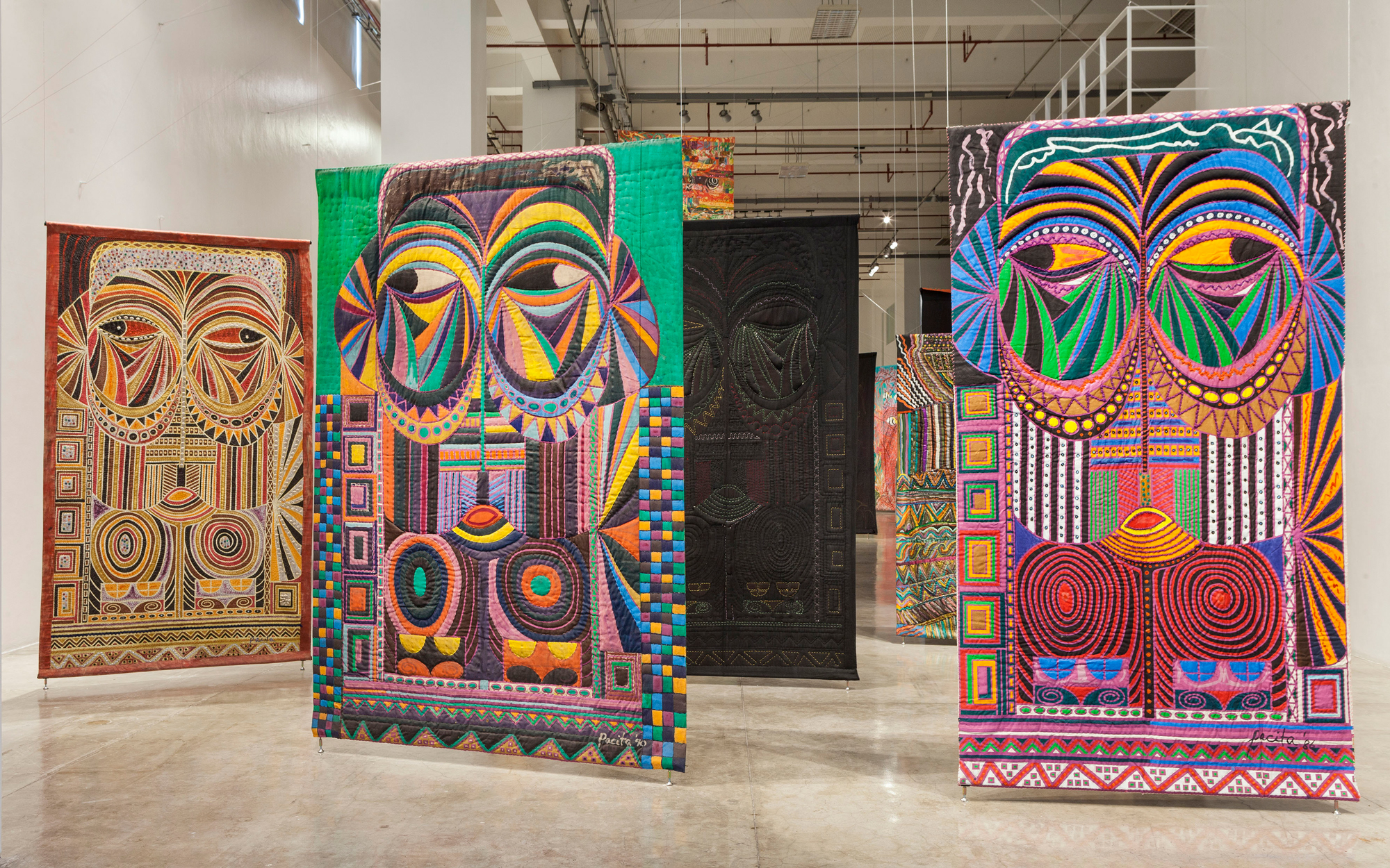 Large gallery space with hanging colorful textile works with mask-like patterns on front and black backing with some stitching visible