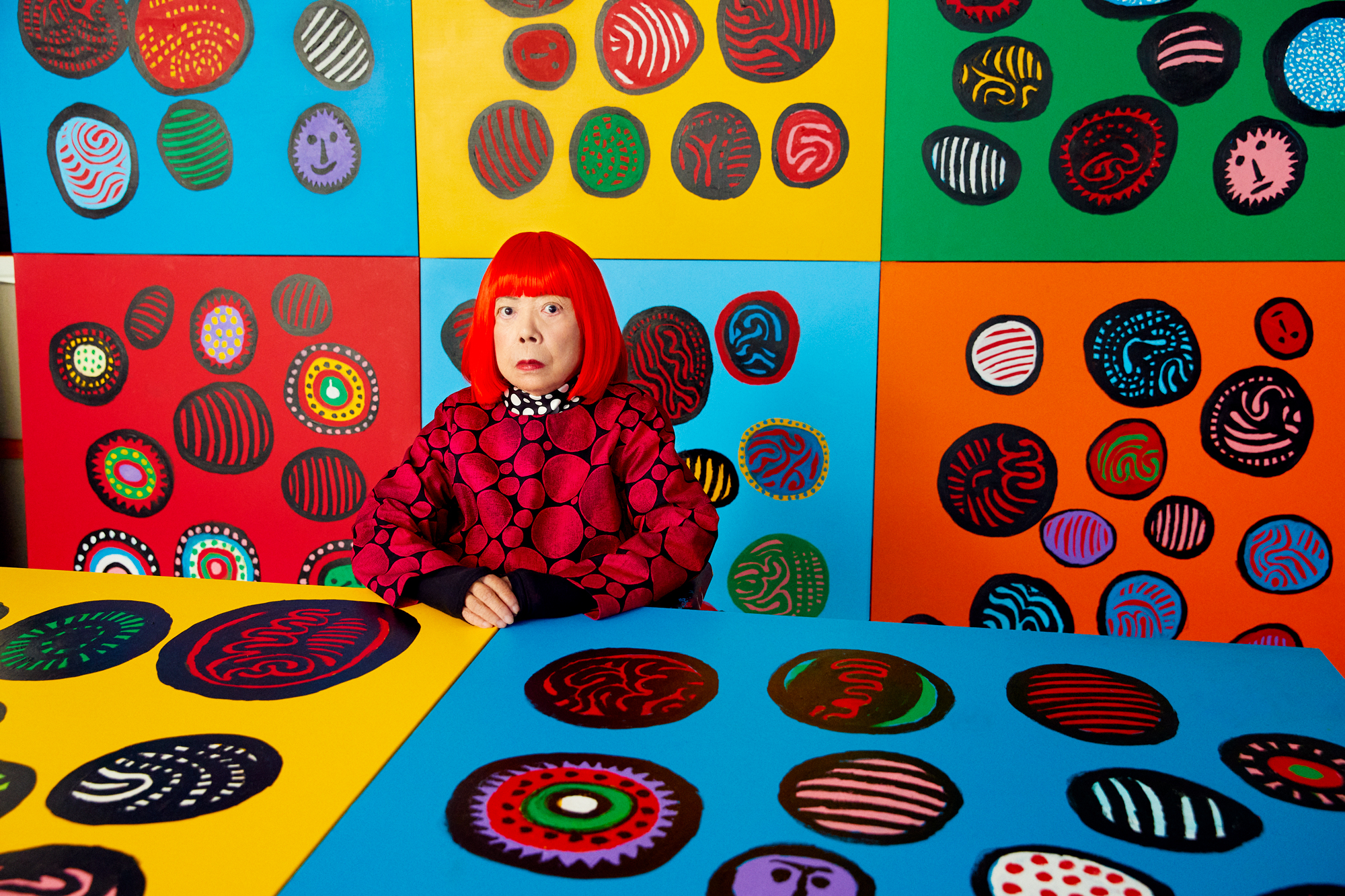 Older woman in bright red bob wig and red jacket, sits surrounded by colorful canvases