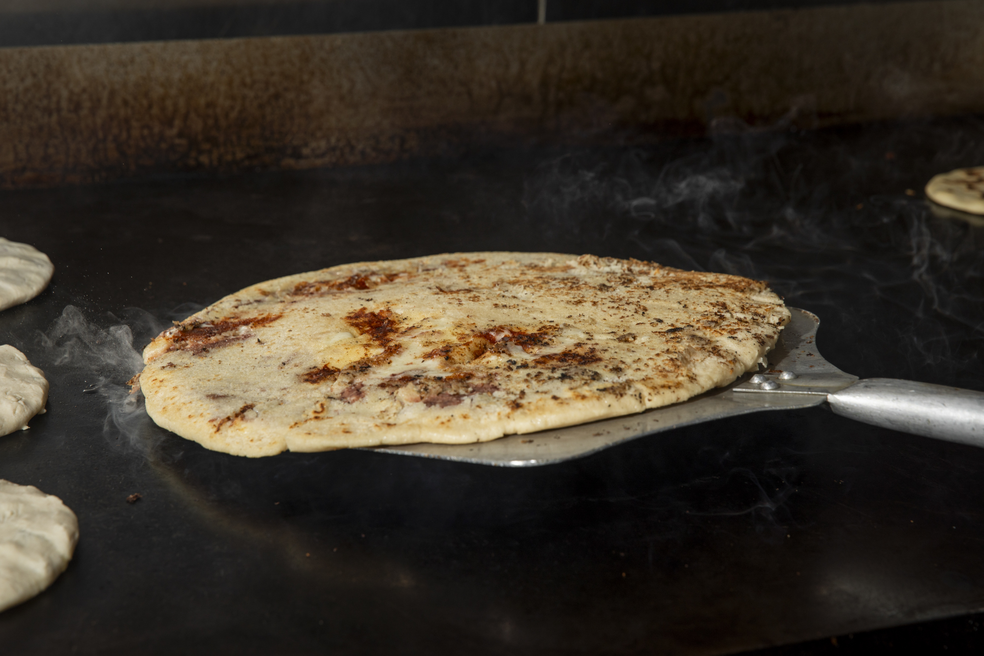 A large pupusa cooking on a griddle is flipped using a pizza peel.