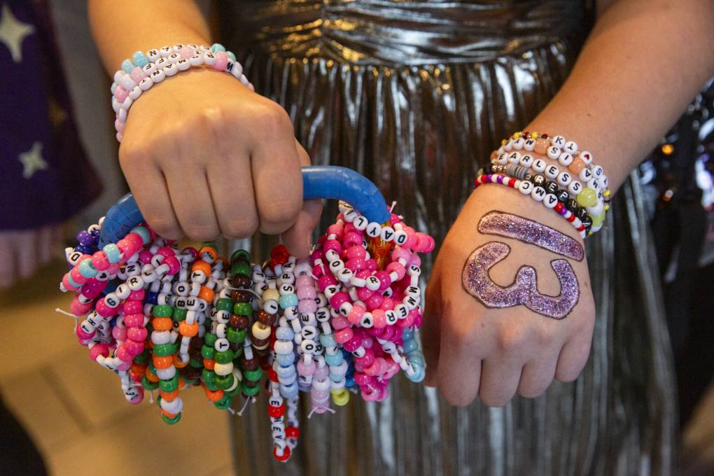 A persons wrists wear eight different bracelets with letters and bright beads on them. The person also holds a carribeaner with dozens more bracelets.