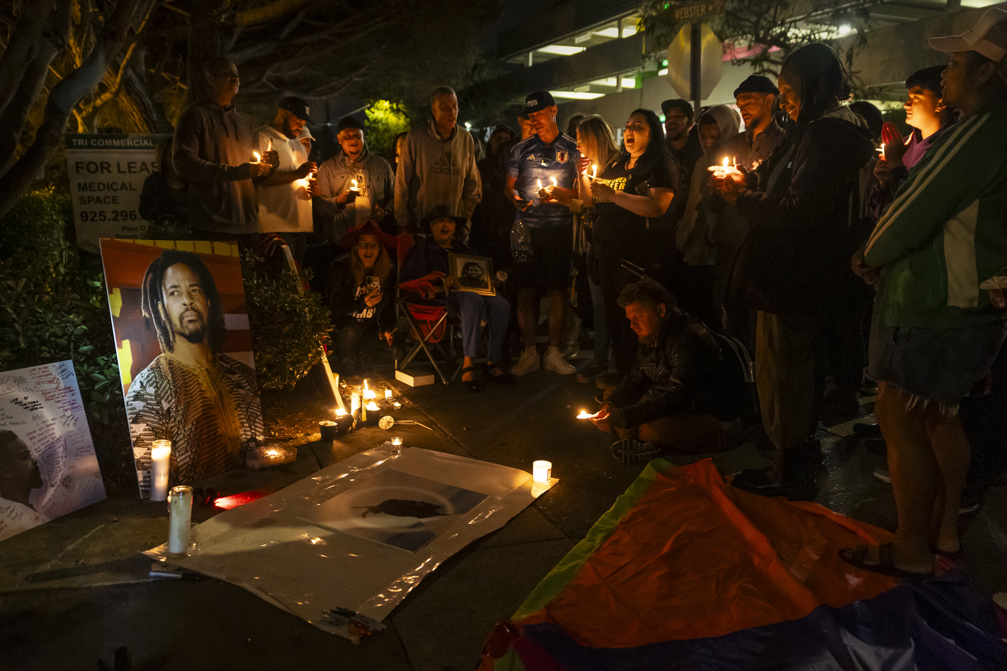 A group of people gather, holding candles and singing around the photo of a person with long hair and a goatee.