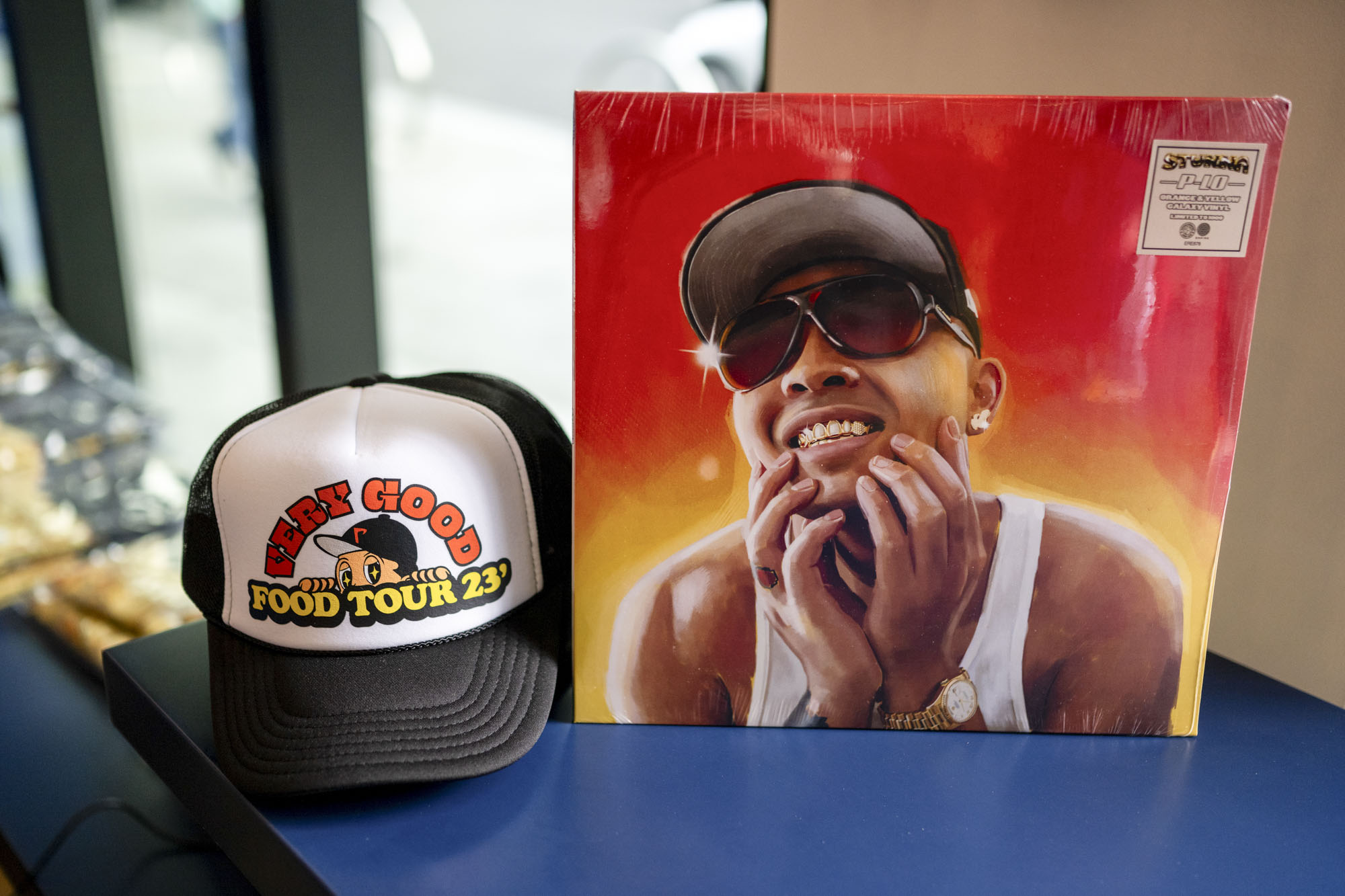 A vinyl album with the photo of a person in a baseball cap on it beside a trucker hat with the words "Very Good Food Tour 23'" written on it.