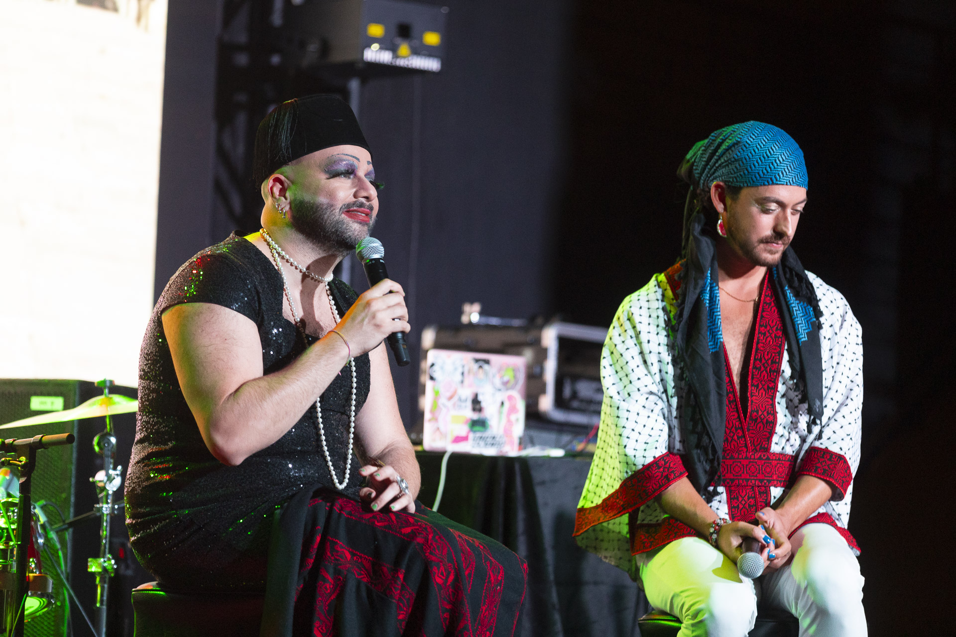 Two Palestinian drag performers wearing feminine clothing and makeup speak on stage.