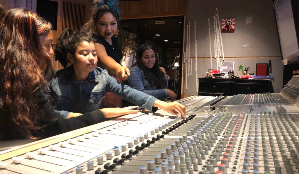 four young women smile while adjusting knobs on a sound board in a studio