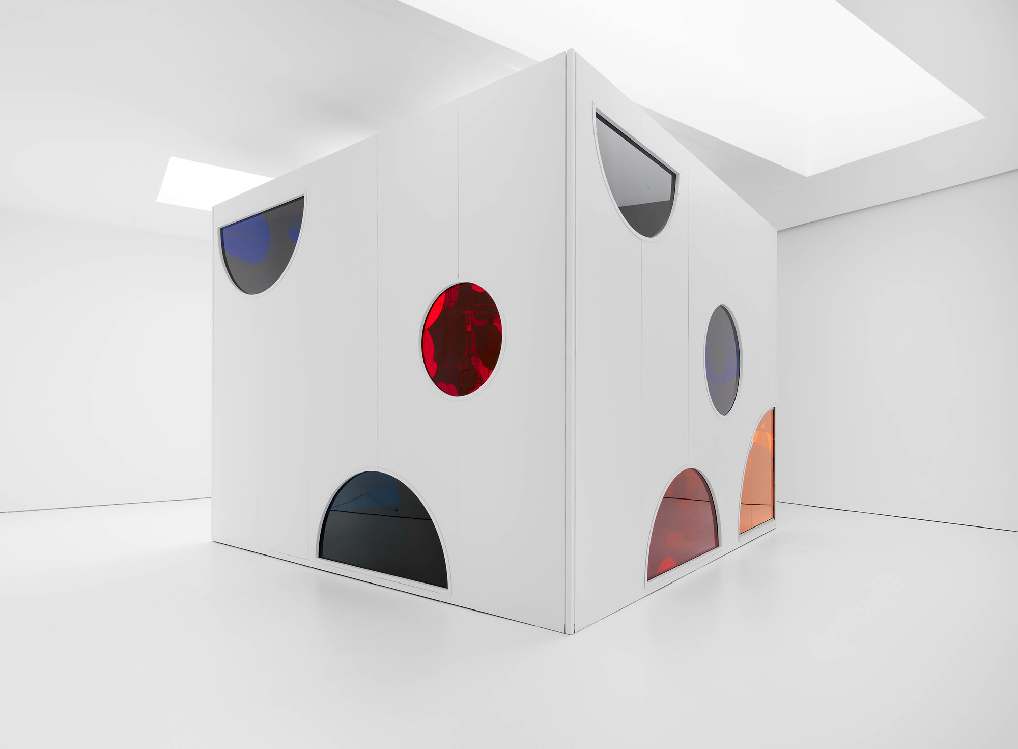 White room with a large white cube at center, dotted by colorful circular windows