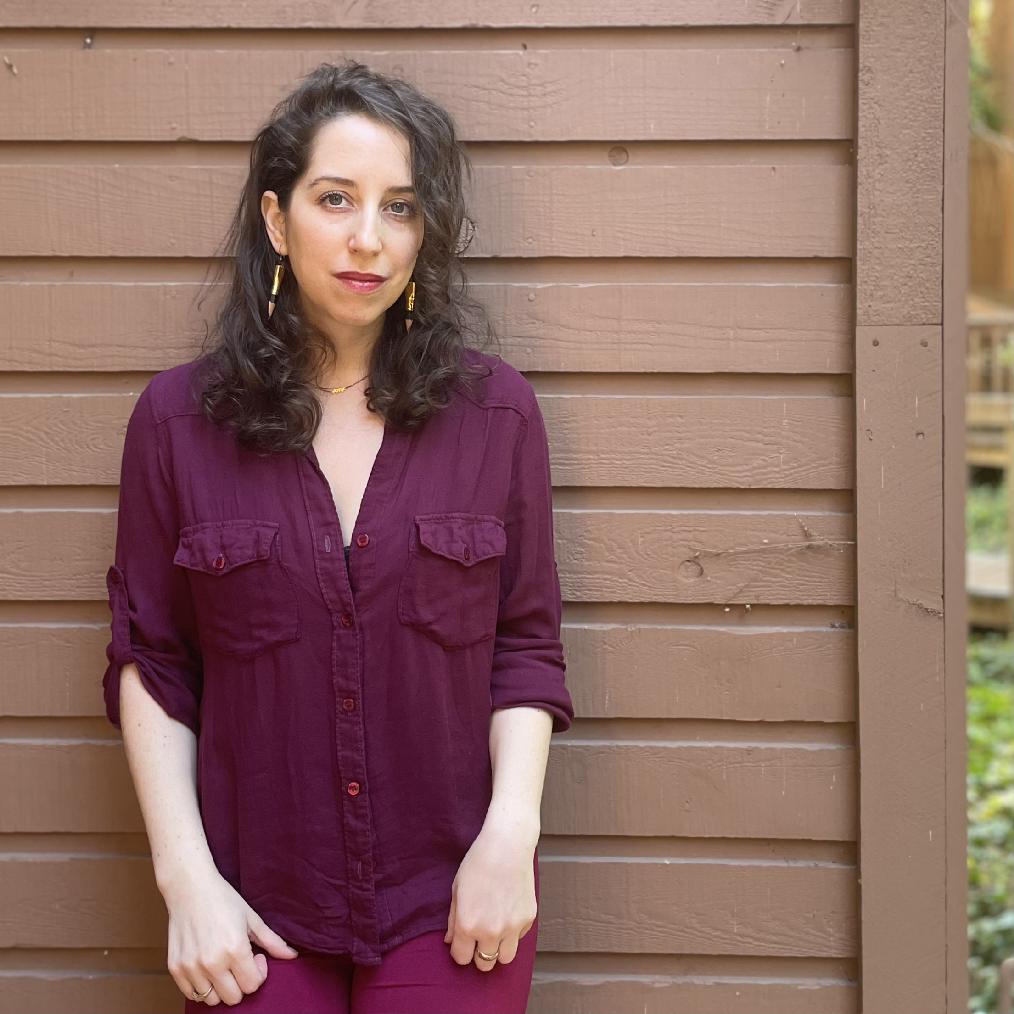 White woman with brown hair in dark purple shirt leans against wooden wall