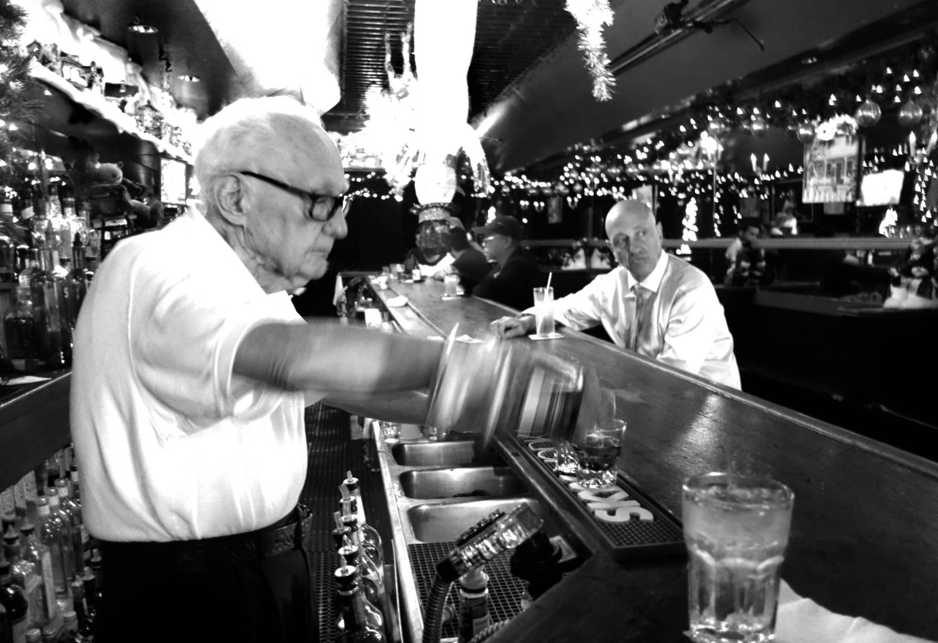 A senior man with white hair and receding hairline pours a drink from behind a small, old-fashioned bar. He is wearing a white short-sleeved shirt and black pants. The bar is full of Christmas lights and lined with mirrors. There are three or four patrons seated at the bar.