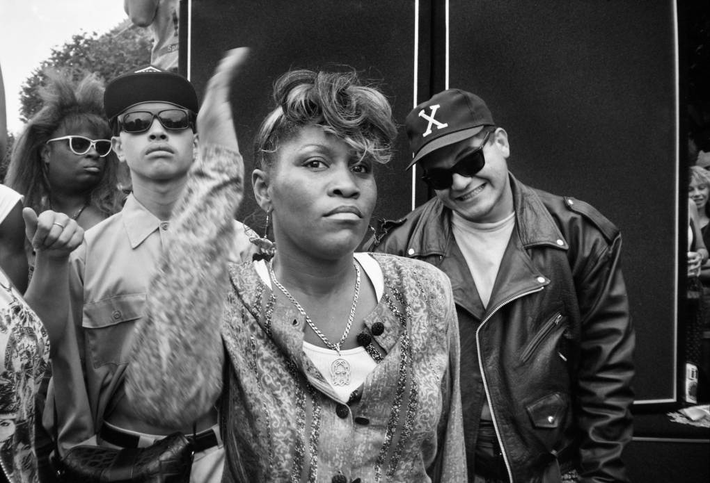 A young Black woman wearing a gold chain, dangling earrings and casual sweater, gestures, putting her right arm at an angle next to her. Behind her on the right stands a grinning, young Latino man wearing a leather jacket and a baseball cap with an X on it. Behind her on the left is a young Black man wearing a work shirt buttoned all the way up, with a baseball cap and sunglasses. Behind them all is a Black person of undetermined gender wearing white sunglasses and backcombed hair.