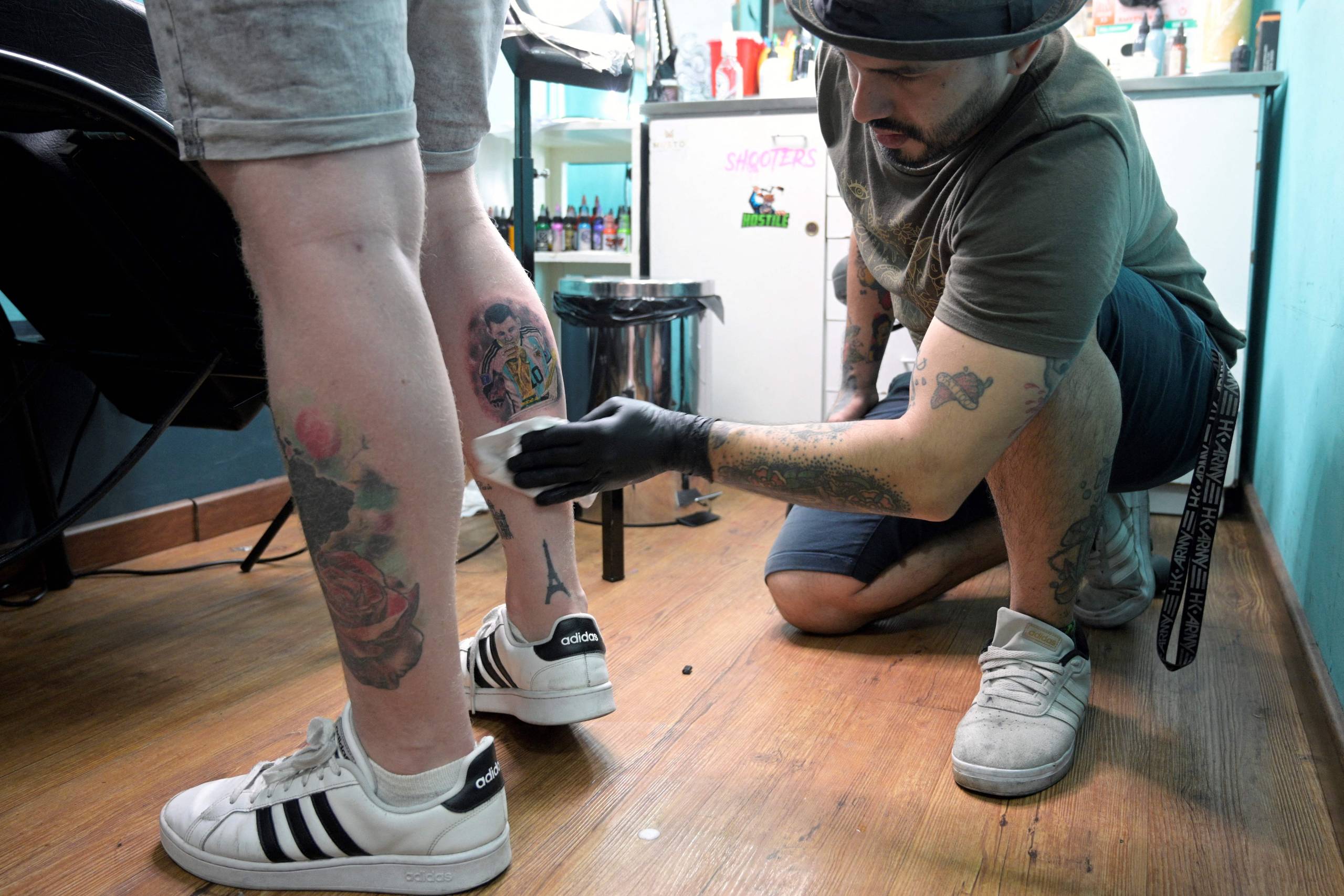 A tattoo artist crouches with one knee on the floor to wipe down a new tattoo on the back of a man's leg.