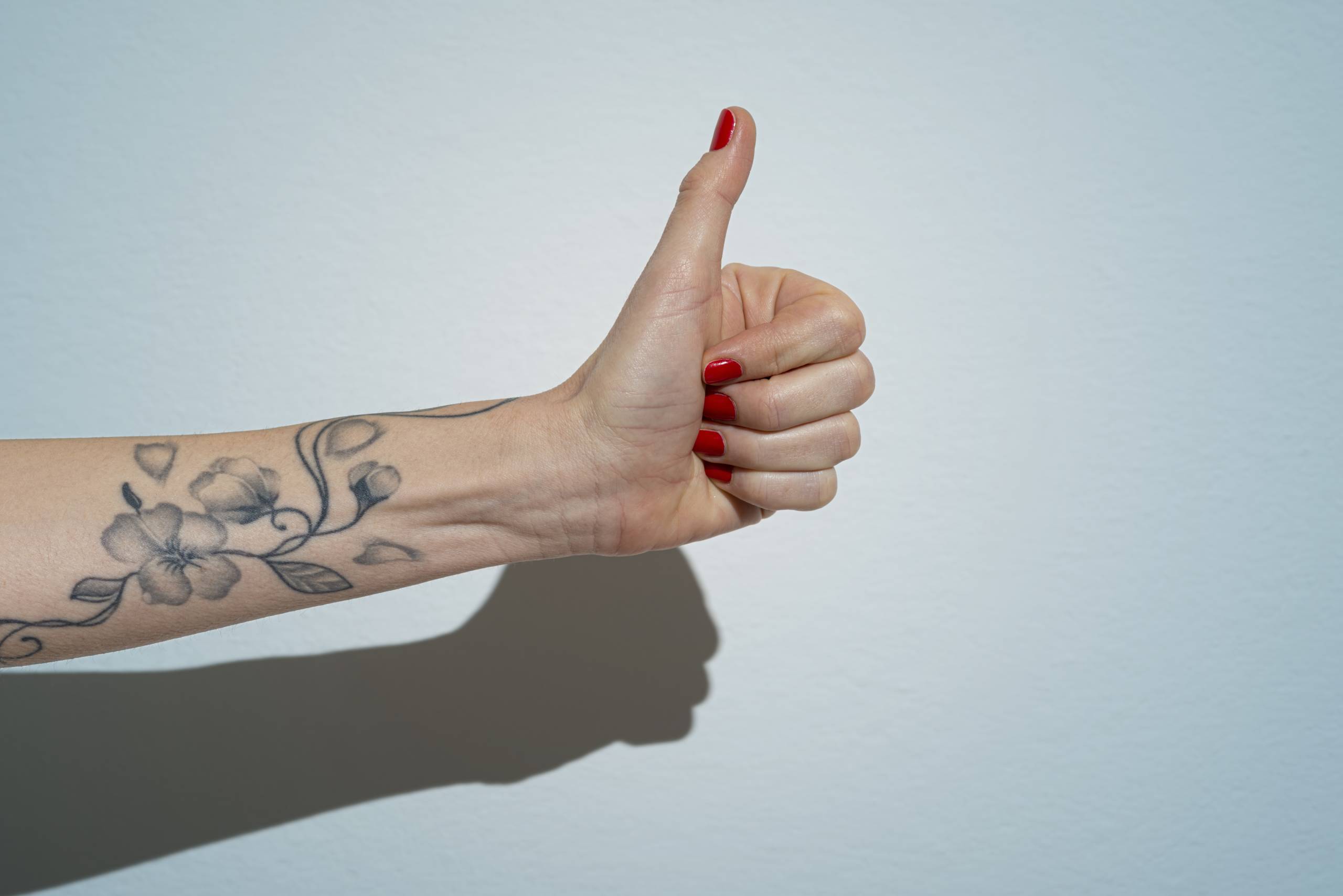 Woman's hand making thumbs up sign against a light blue wall. She has red nail polish and delicate flowers tattooed on her forearm.