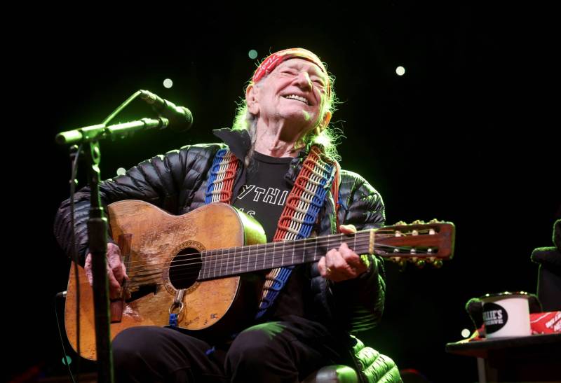 An elderly man sits on stage behind a microphone, playing an acoustic guitar and smiling broadly as he looks out over the audience.