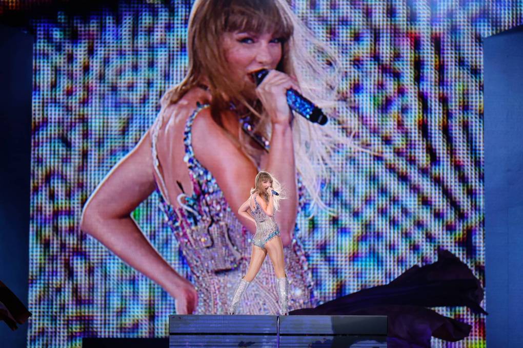 Taylor Swift in sequinned leotard and knee high boots stands on a platform on stage, directly in front of a big screen broadcasting her live image. She is holding a microphone to her mouth.