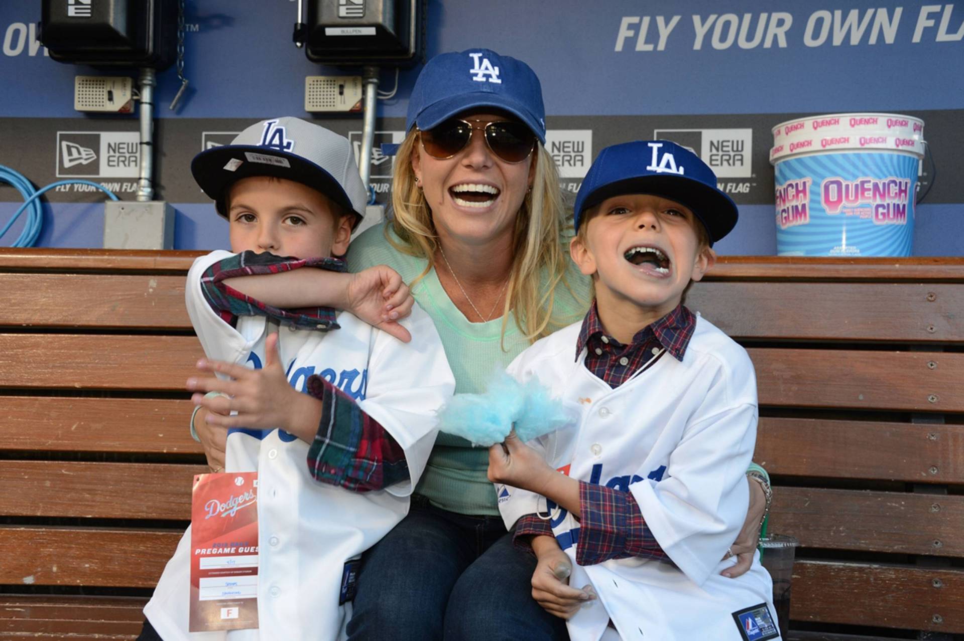 A laughing young woman wearing a green sweater and an blue LA baseball cap embraces two young boys. The children are wearing LA Dodgers jerseys and baseball caps.