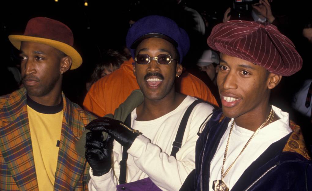 Three young Black men wearing vibrant 1990s-era clothing and hats smile in a crowded room.