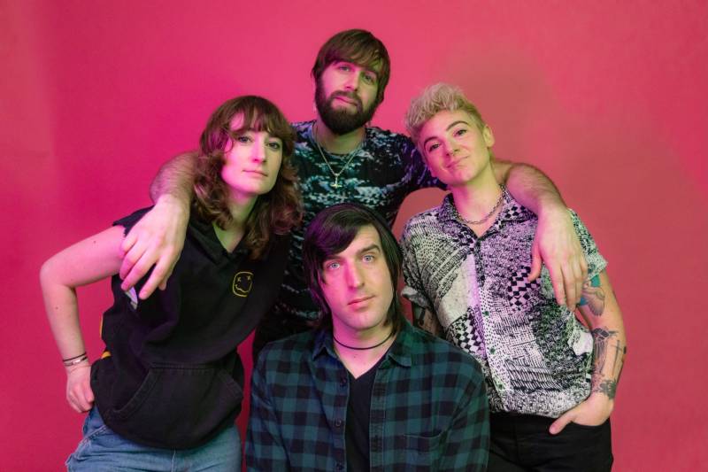 a four-person band of youngish white people in colorful shirts pose for a band portrait against a pink background