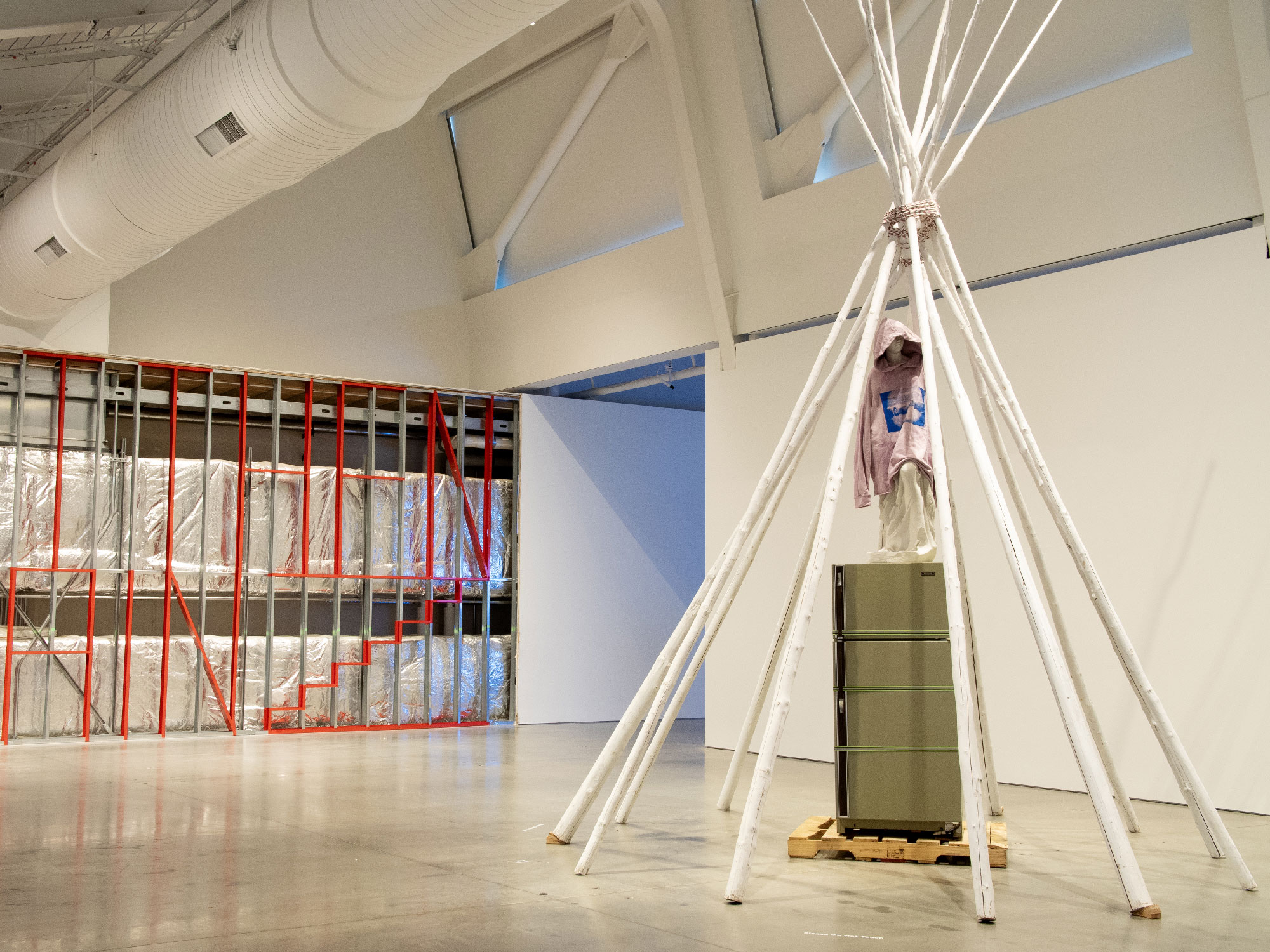 A white-pole tepee sculpture in a gallery space, with a wall behind partially demolished, red paint on the metal beams spells out words