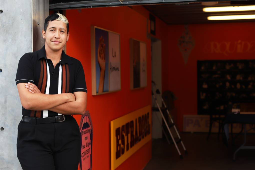 Artist Marcel Pardo Ariza, a Latine trans masculine person in their 30s, stands before an open garage door. Parts of their installation of photography and archives are visible in the background.