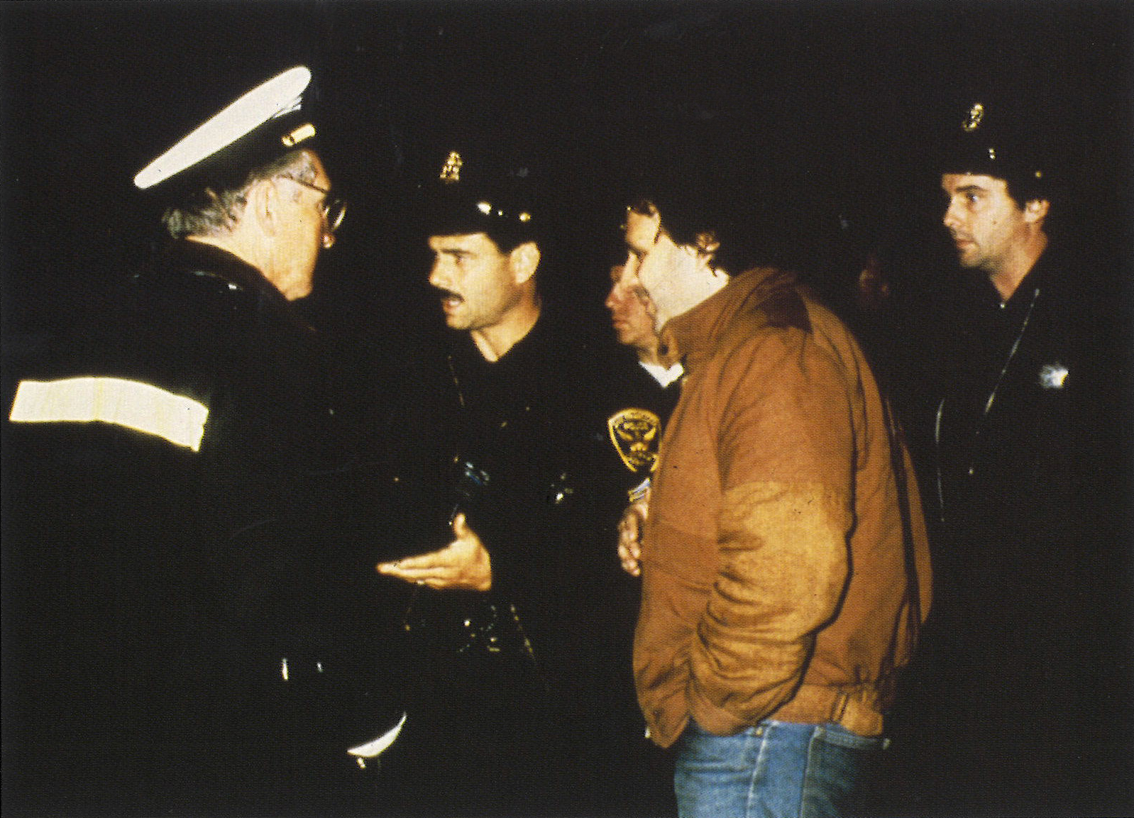 Photograph of four  police and firefighters and a man in a tan jacket against a black background