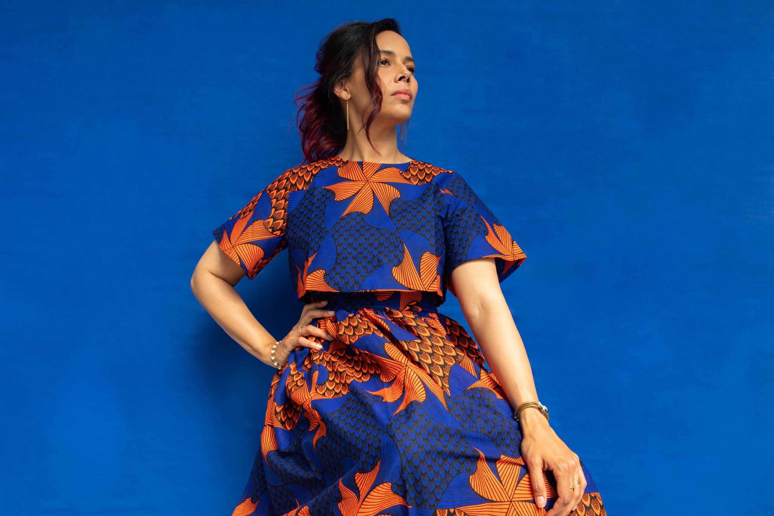 a woman with light brown skin and dark brown hair poses in a blue and orange dress against a blue backdrop