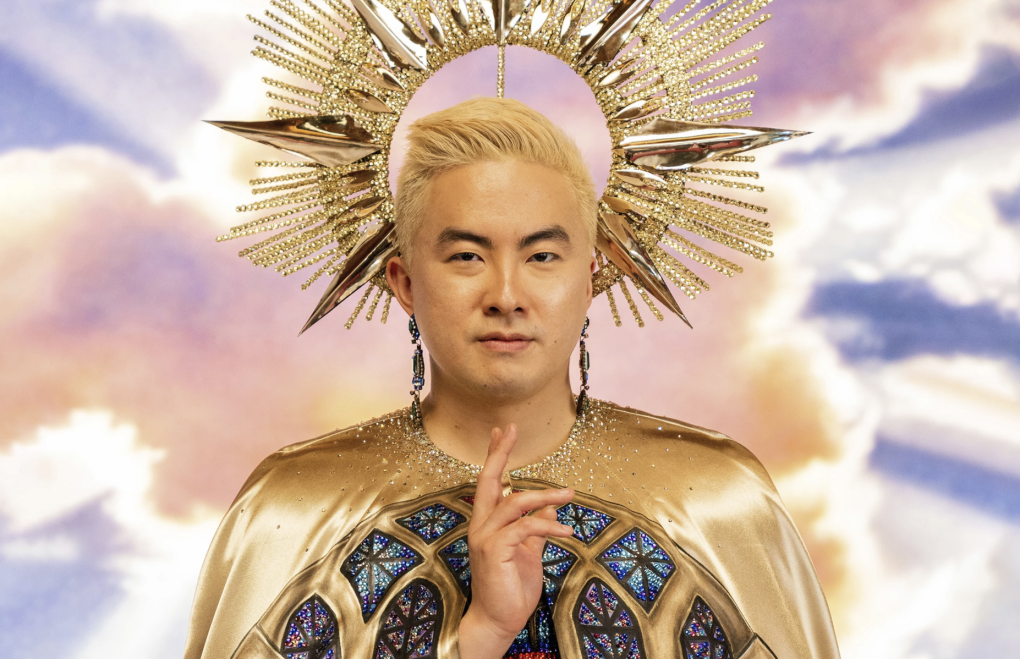 A young Asian man with blonde hair, wearing sumptuous gold robes stands before a backdrop of pink and white clouds, wearing an elaborate gold headpiece.