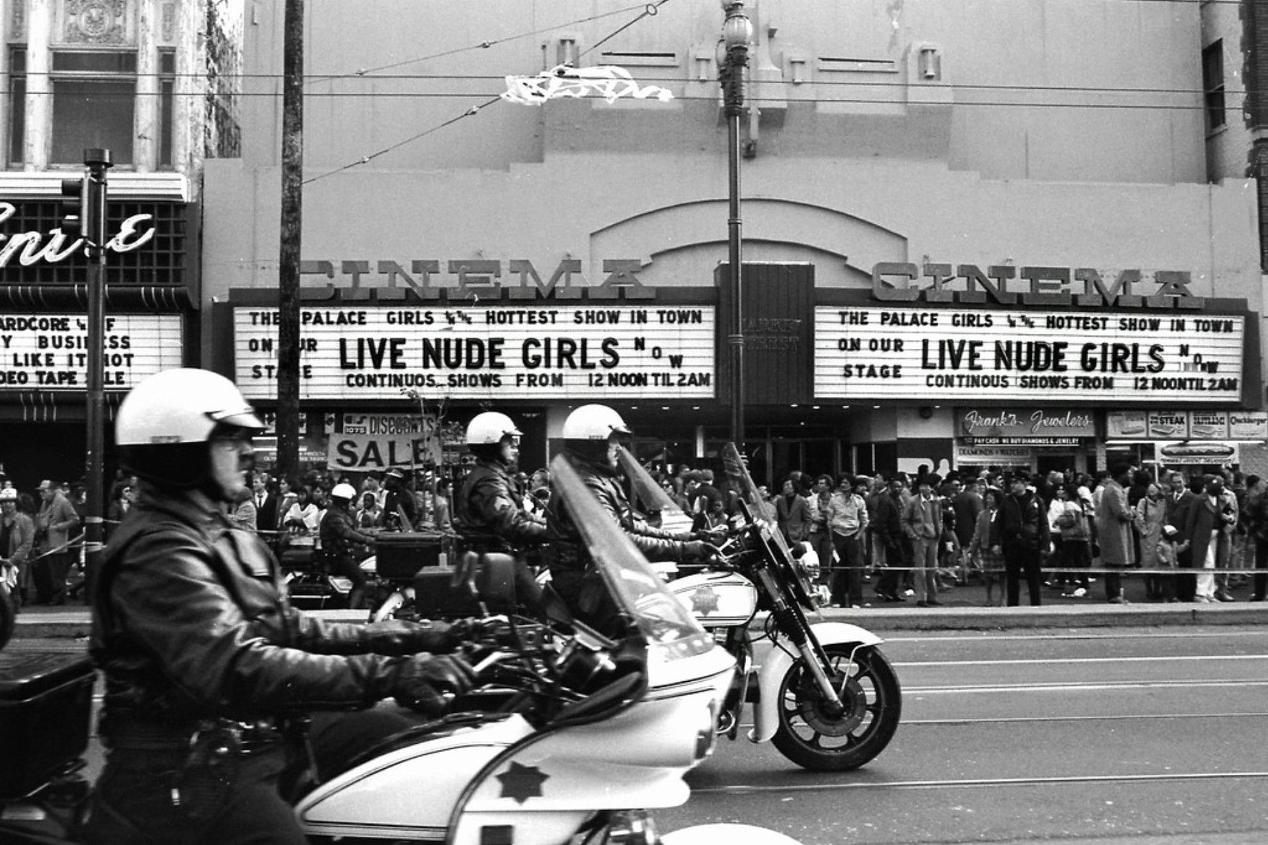 A line of motorcycle cops block off the entire street. Barricades hold back crowds on the sidewalk behind them. A cinema promises LIVE NUDE GIRLS from its marquee.