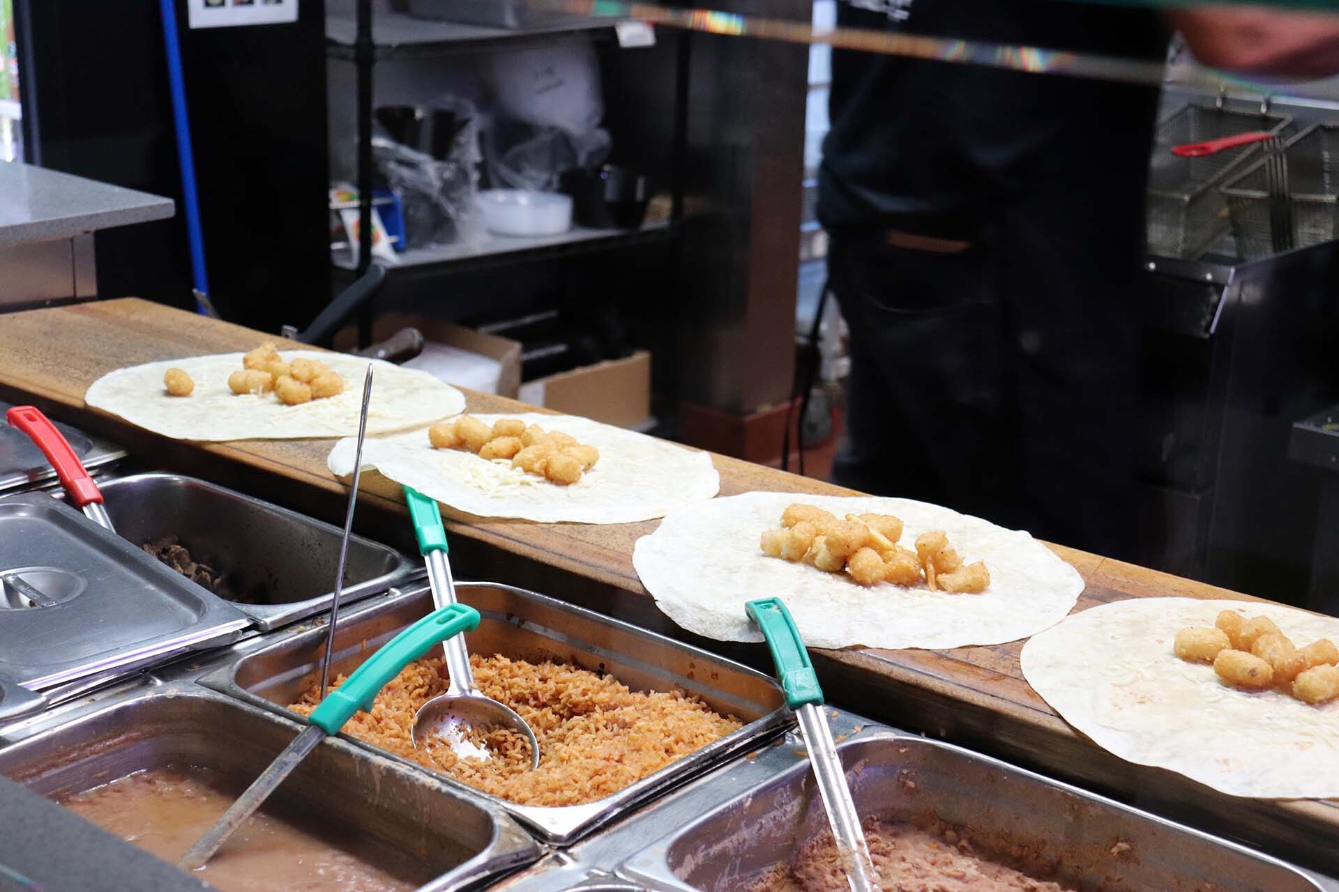 inside a taqueria's kitchen, four burritos are being prepared with tater tots as a prominent ingredient