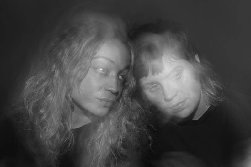 A blurry, black-and-white photo of two young musicians, one female and one nonbinary, leaning their heads together.