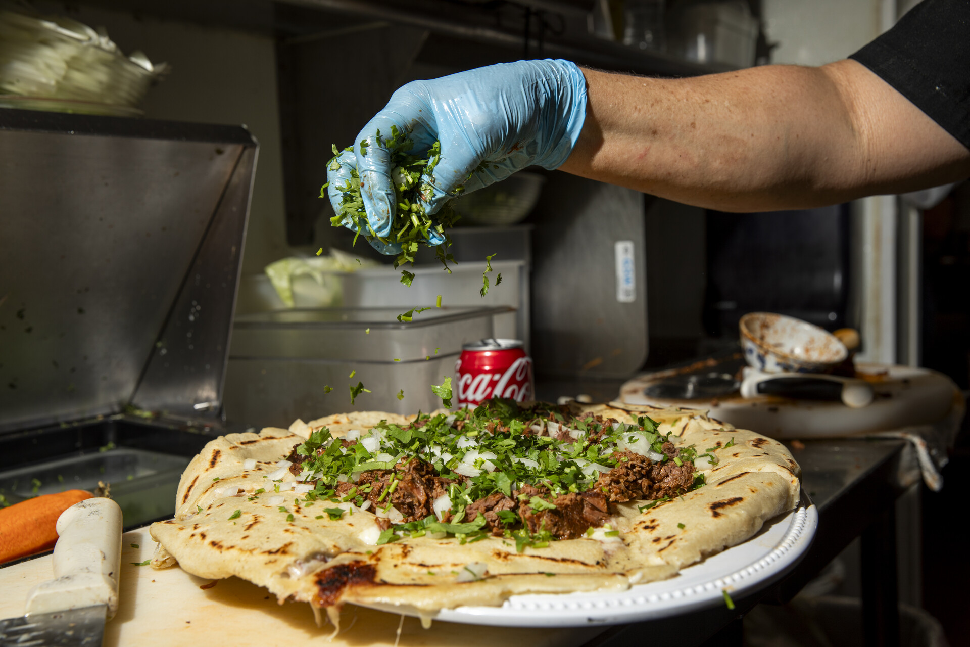 Rubber gloved hand sprinkles chopped cilantro onto an oversized pupusa topped the meat.