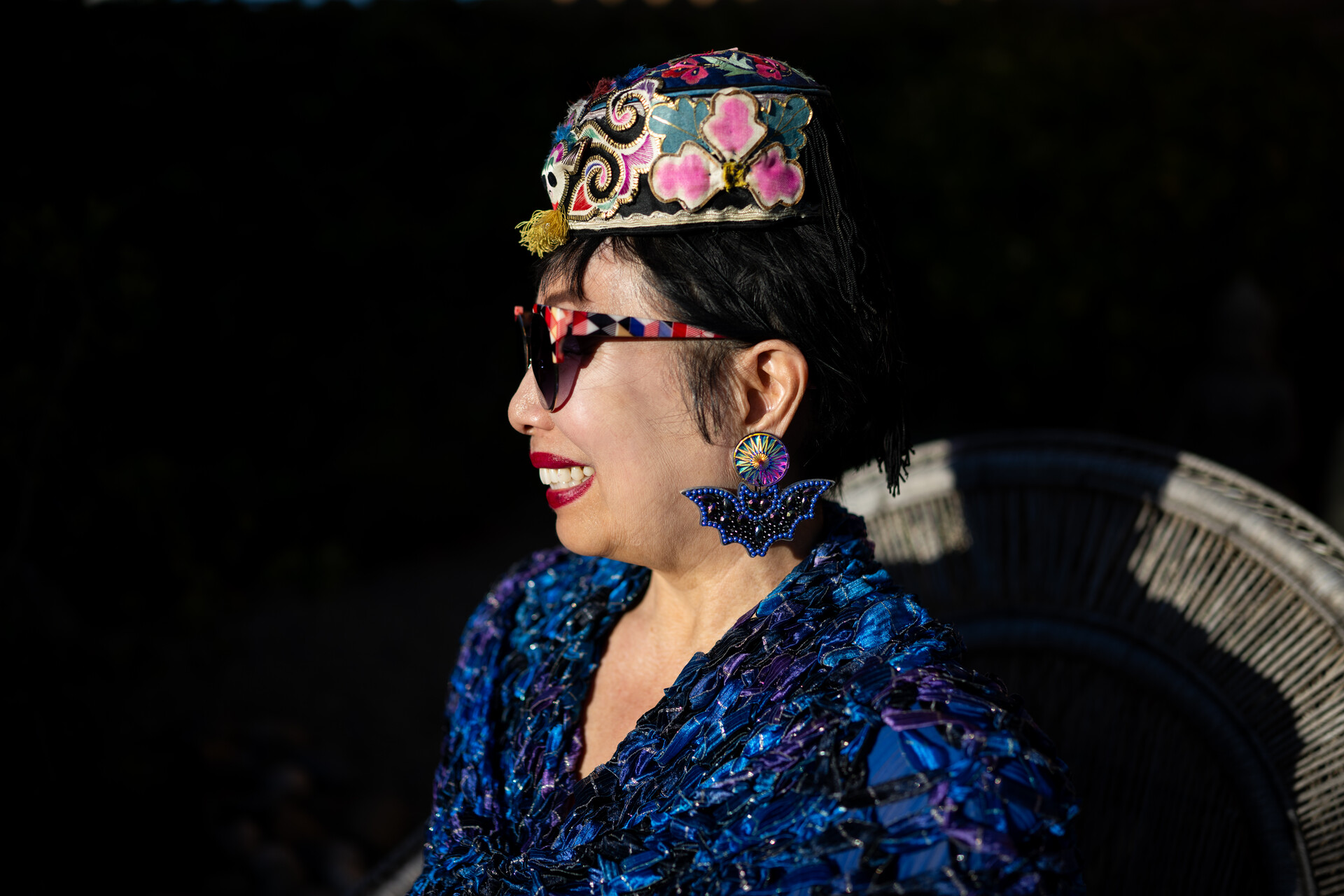 Asian woman in sunglasses, embroidered hat and bat-shaped earrings smiles in profile