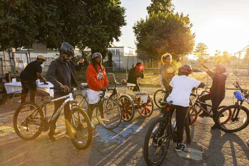 A group of adults and children hang out on bikes at a park.