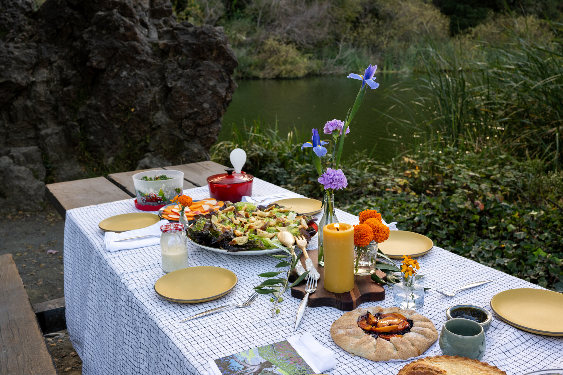 A colorful outdoor table setting with a black-and-white checkered tablecloth, candles and fresh flowers — plus a fruit galette and big platter of salad.