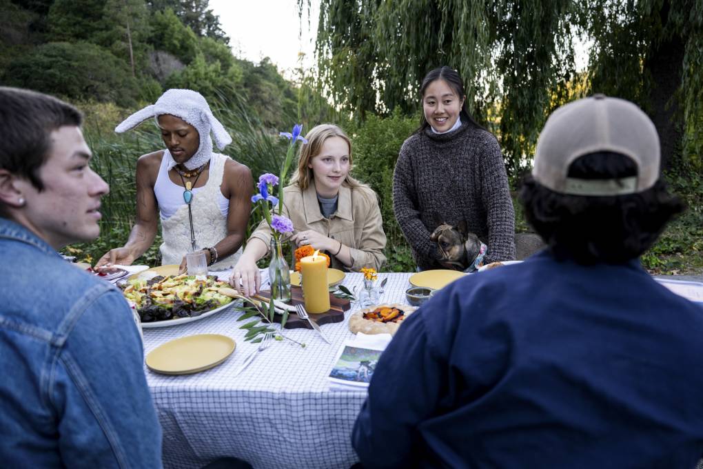 A group of people talk around a picnic table set with a table cloth and a meal.