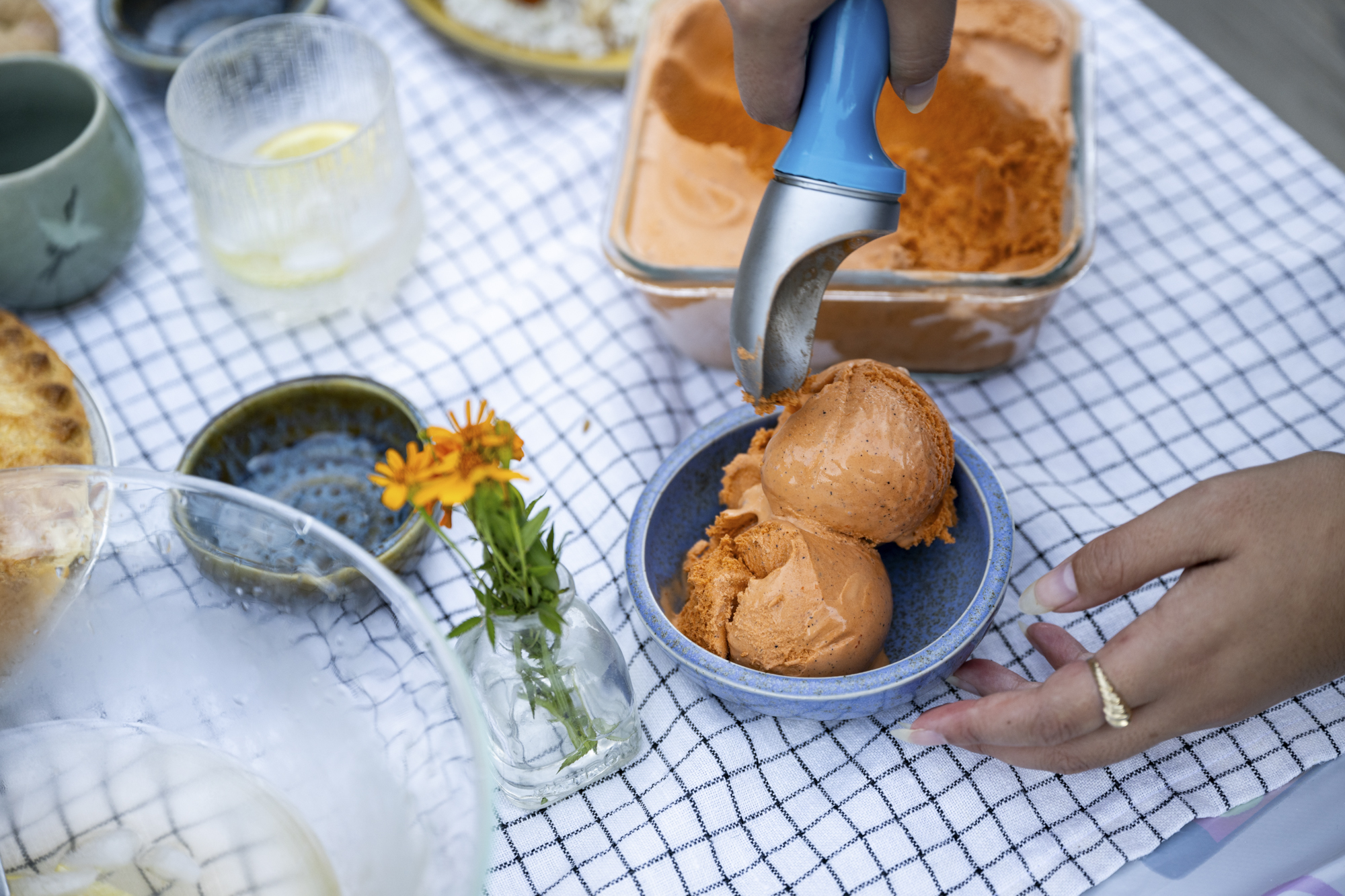 A person serves themselves brownish orange-colored ice cream at a picnic table set with a tablecloth and a meal.