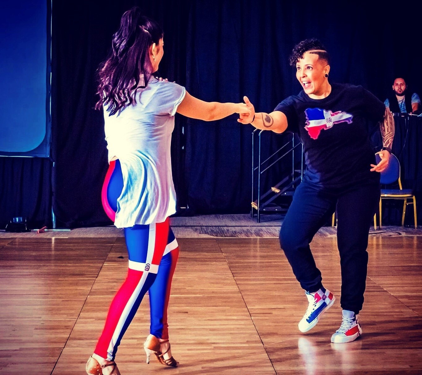 A woman in multi-colored leggings faces away from the camera, while dancing and holding the hand of a woman facing the camera in a black top and black jeans.
