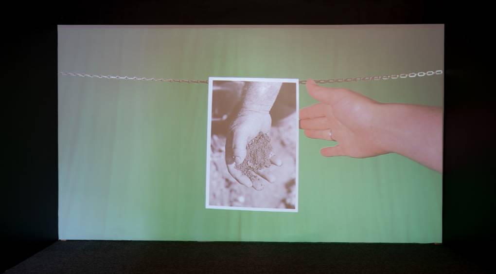 A white hand reaches out to touch a black and white photograph of a hand holding dirt against a green background