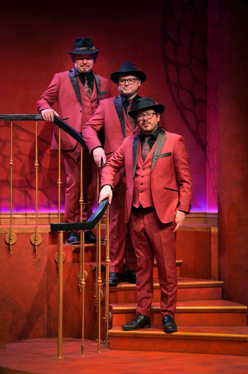 three men in a jazz ensemble dressed in red suits and black bowler hats stand on a staircase on a stage