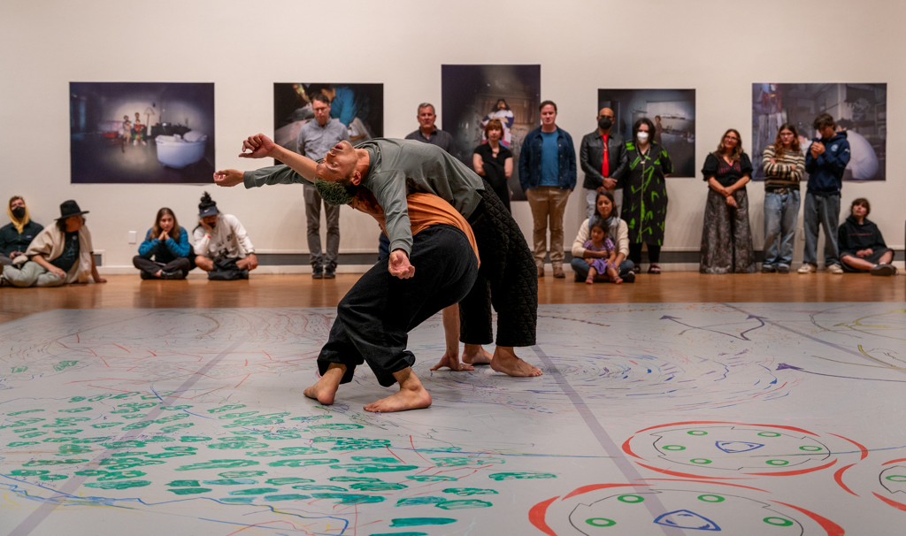 Two dancers lean against each other while audience watches in gallery, floor is covered with colorful patterns