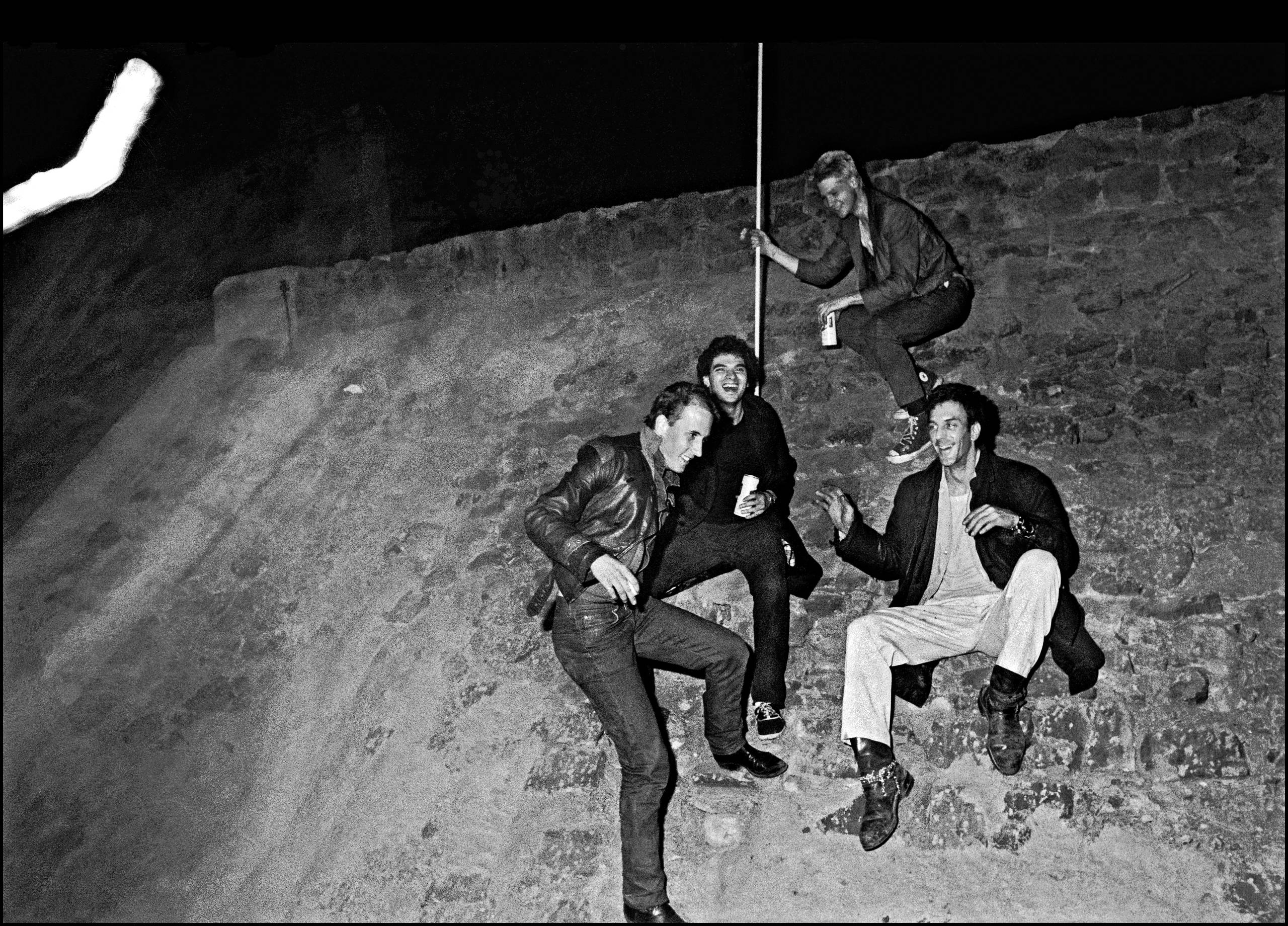 A group of four young men clamber down a very steep wall, laughing and at strange angles. One of them is holding a pool cue. Two of them are holding cans of beer.