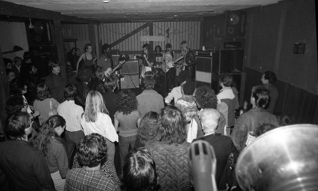 A small, dank room with a band playing at the back of the room and a small audience watching, viewed from behind.