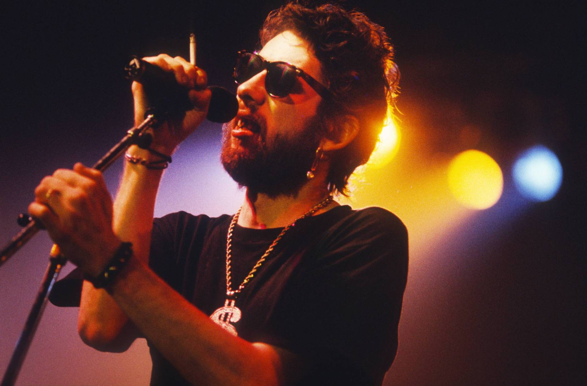 A thin white man with a beard, wearing sunglasses clutches a microphone on a stand, as he sings into it. He is backlit by stage lights.