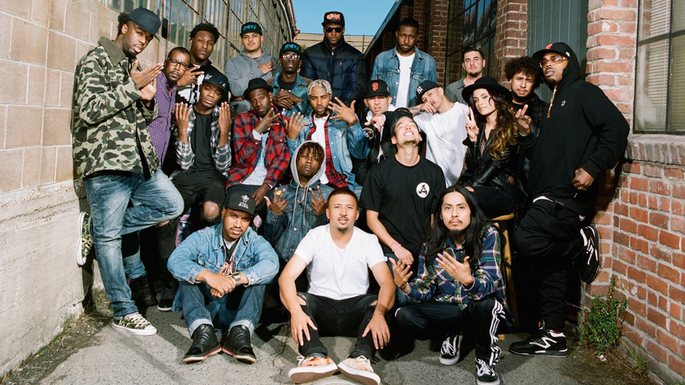 A large crew of rappers and producers poses in an alleyway.