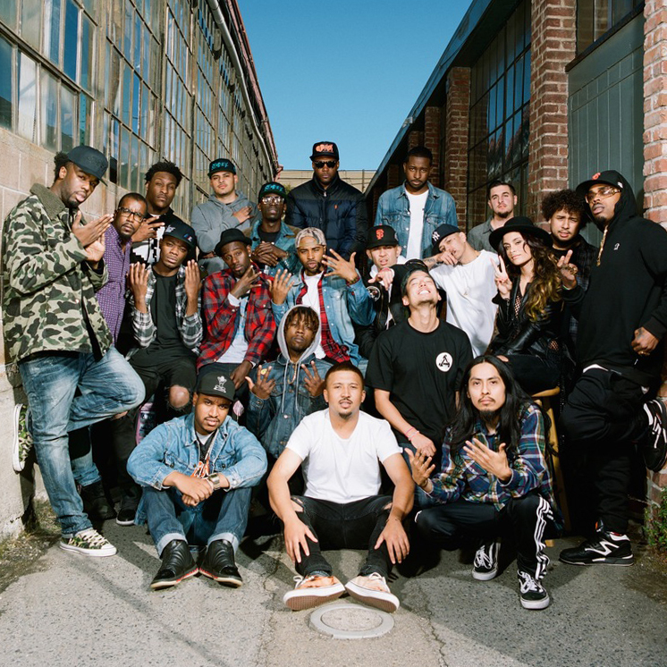 A large crew of rappers and producers poses in an alleyway.