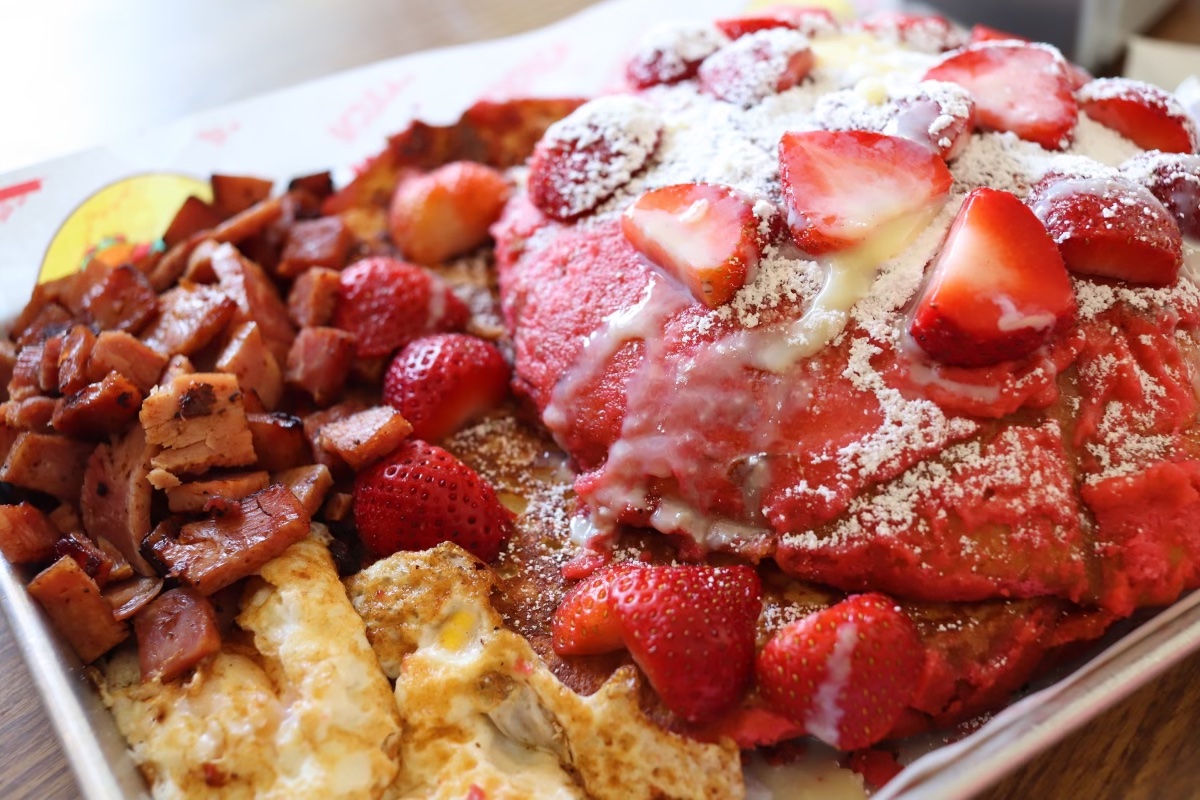 a tray of pink pan dulce french toast is topped with strawberries and served with a hefty side of eggs and ham